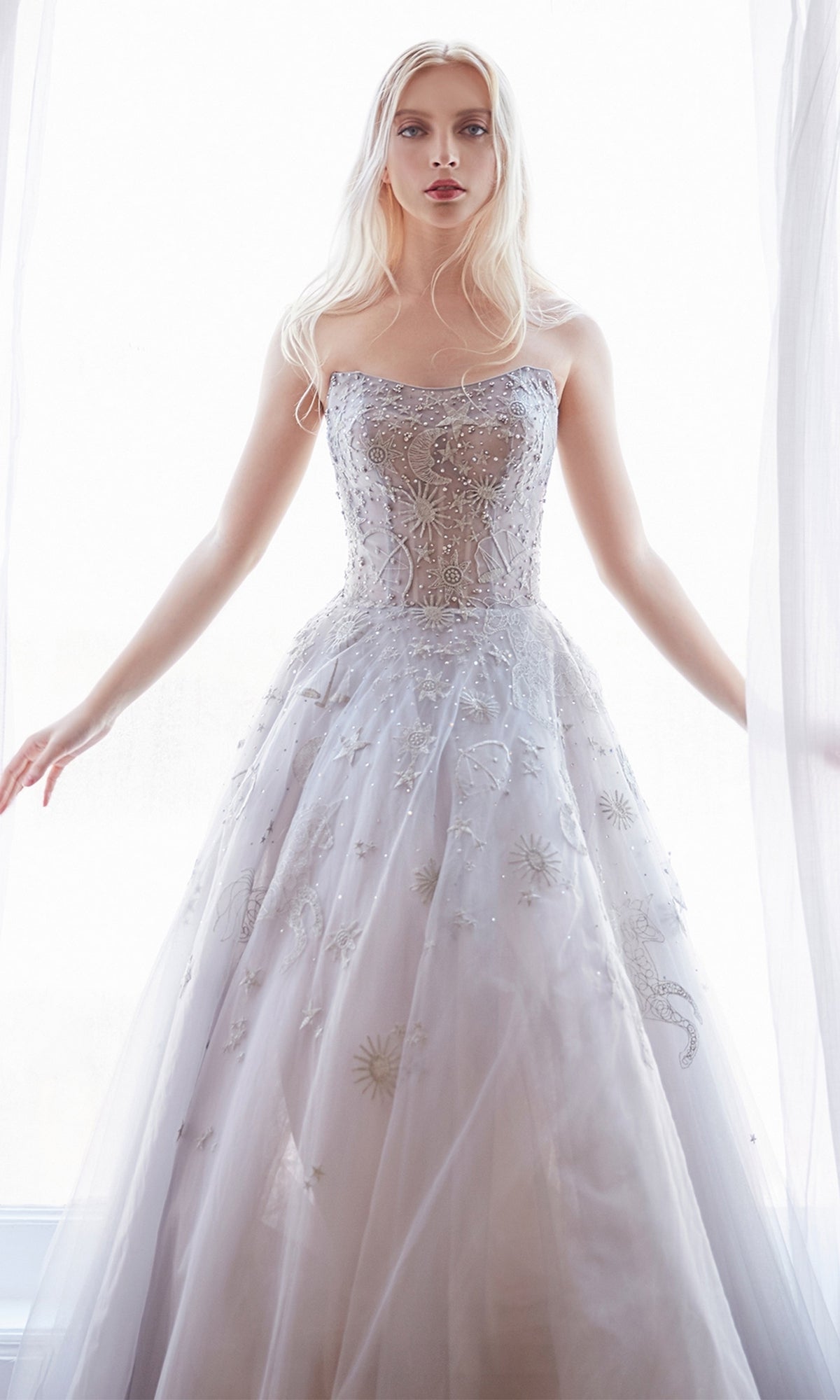 A woman wears the Constellation-Print Strapless Prom Ball Gown A0890, standing with arms slightly extended, framed by sheer curtains and illuminated from behind by natural light.